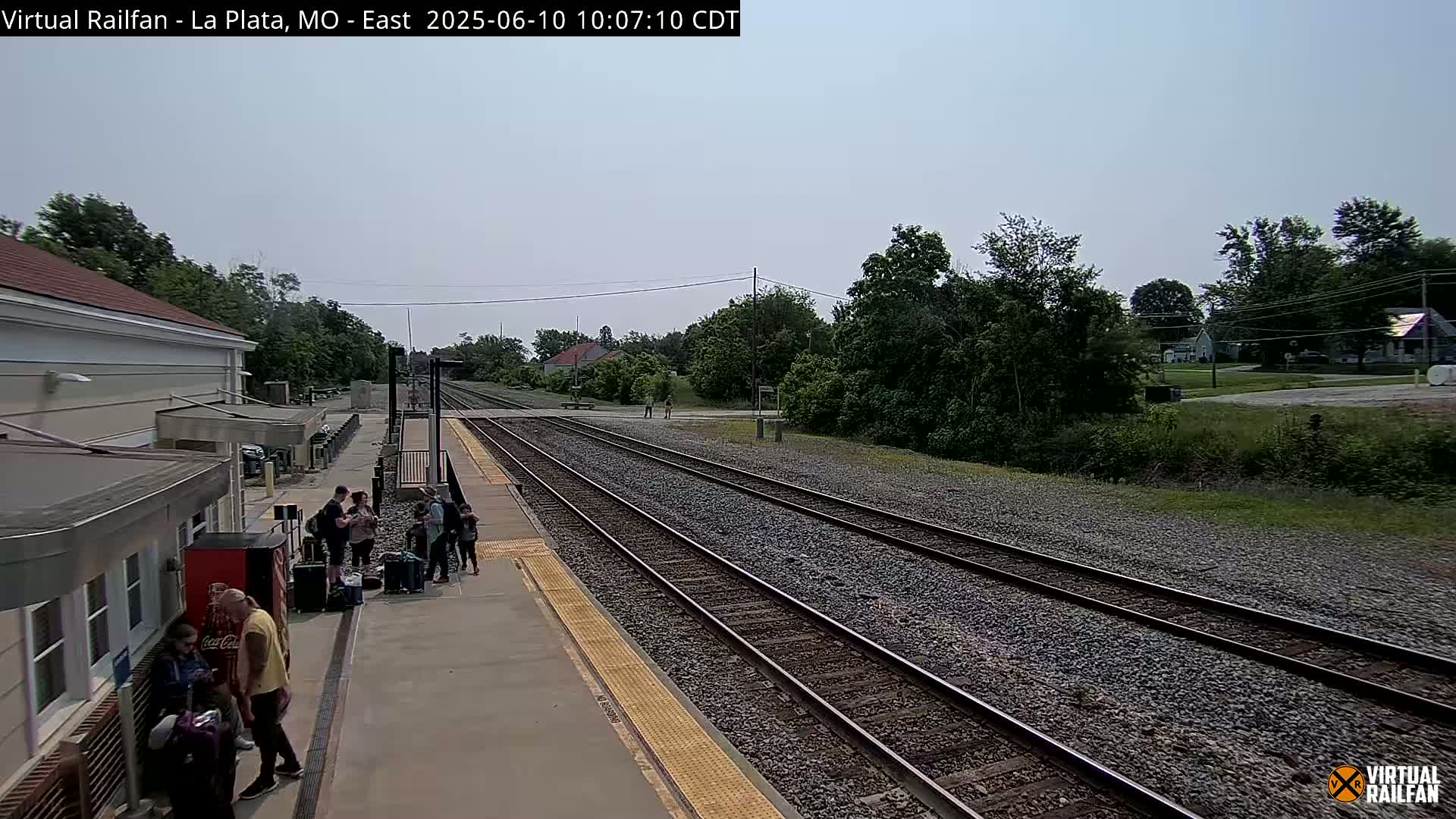 A train station platform with several people waiting near luggage, under a partly cloudy sky, next to multiple train tracks that extend into a wooded area.