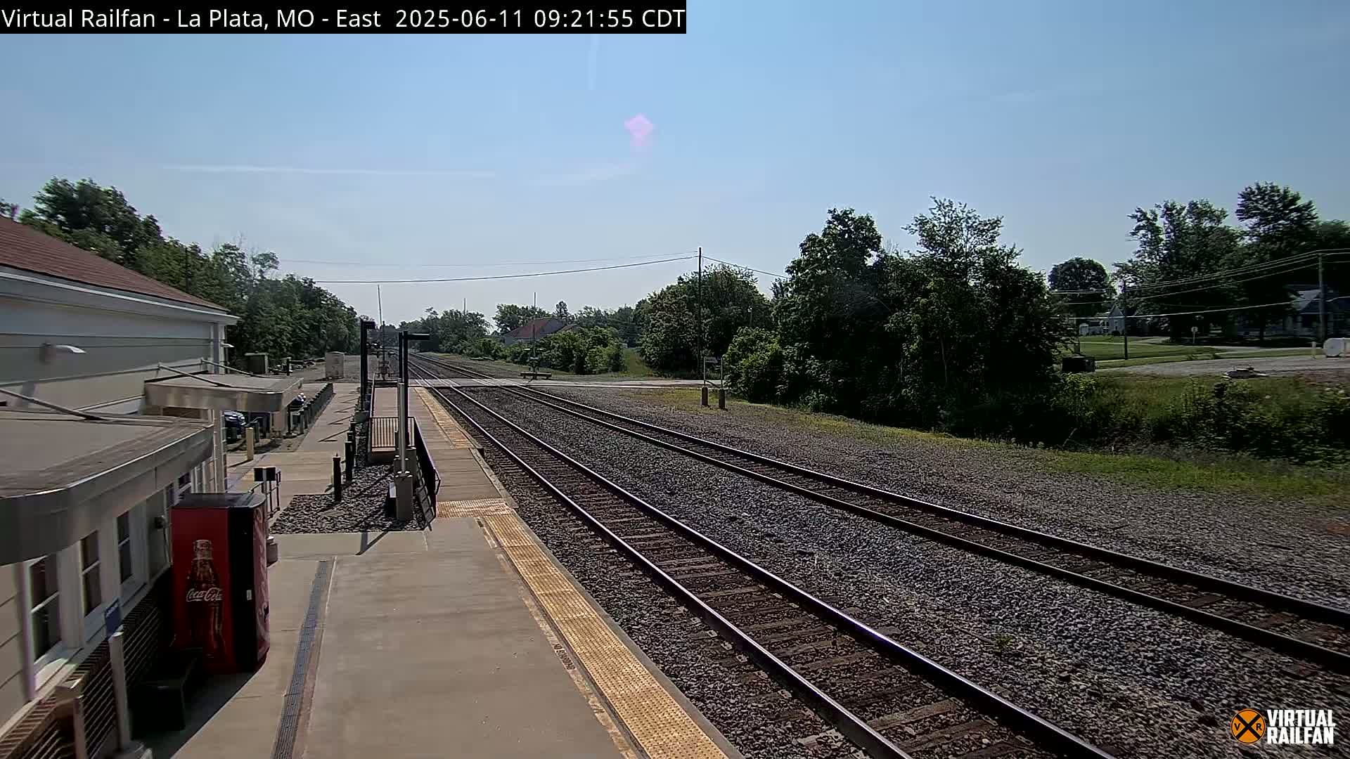A train station platform with multiple train tracks extends into a sunny, clear day, surrounded by green trees and a small town in the distance.