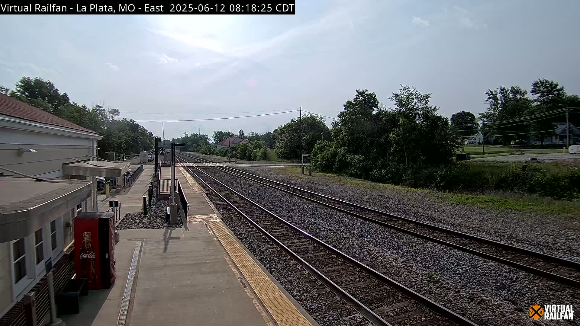 A mostly sunny day view of a train station platform and tracks extending into a tree-lined landscape.