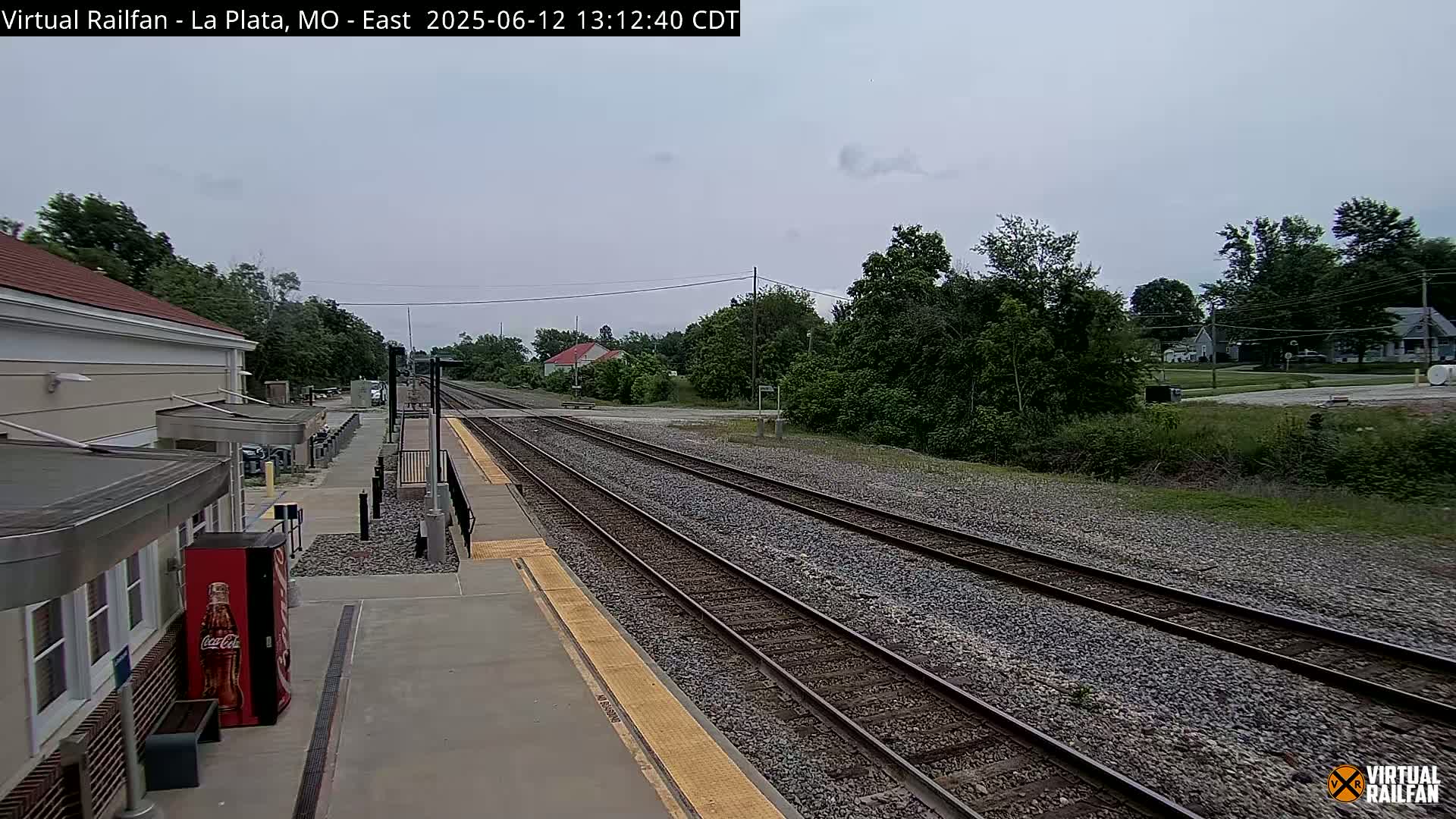 A train station platform with multiple train tracks extending into a landscape of green trees and houses under an overcast sky.