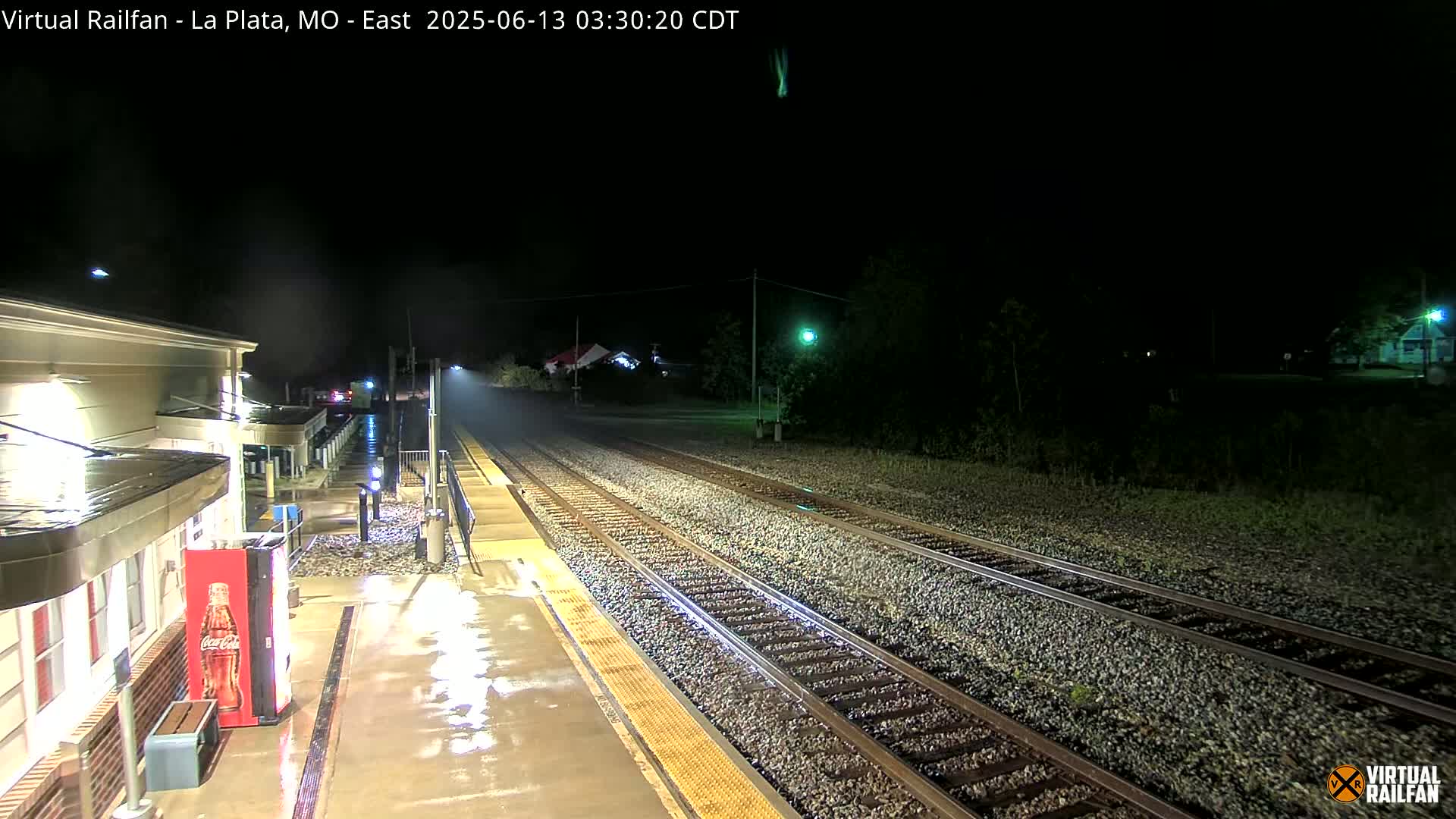 A rain-slicked train platform at night shows empty train tracks extending into the darkness.