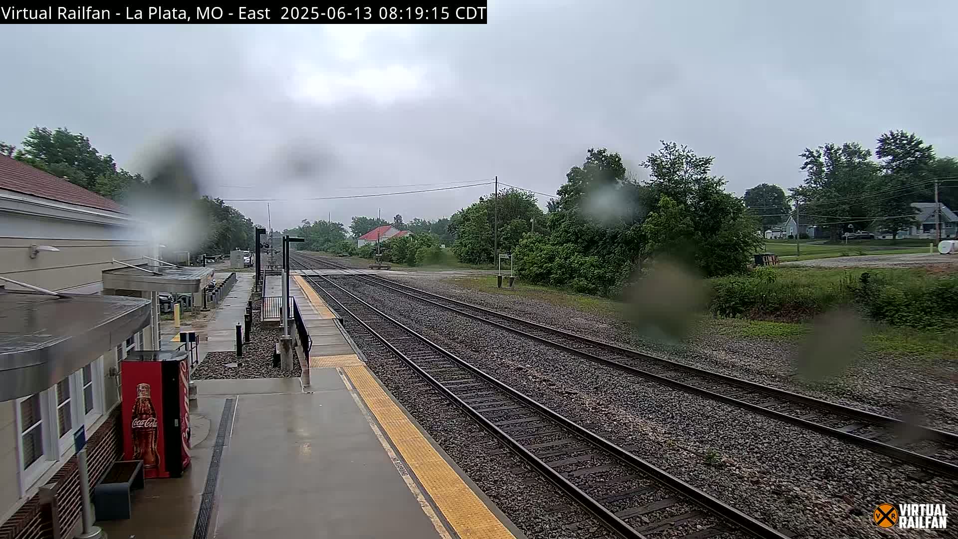 A rain-soaked train station platform with multiple train tracks extending into a verdant landscape under an overcast sky.
