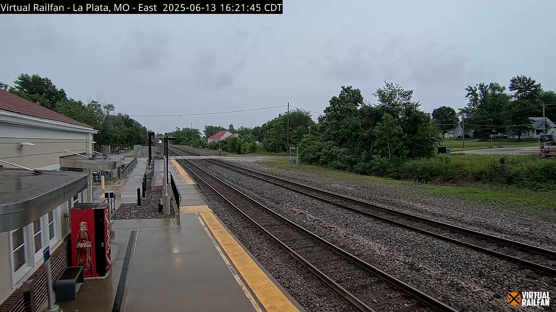 A rain-slicked train platform at a station overlooks multiple train tracks that extend into a landscape of trees and houses under an overcast sky.