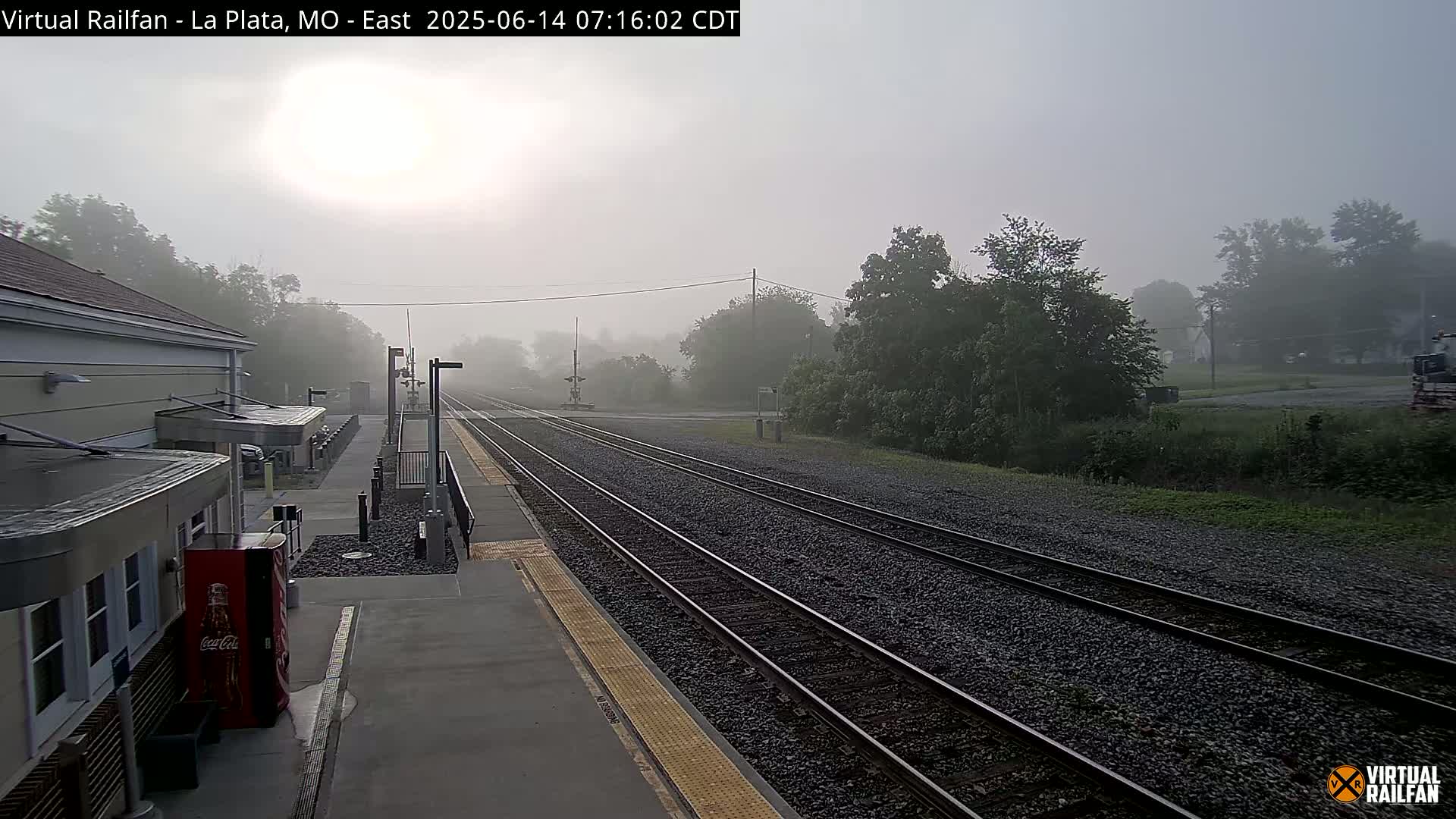 A foggy morning scene at a train station shows multiple train tracks extending into the distance, alongside a building and platform.
