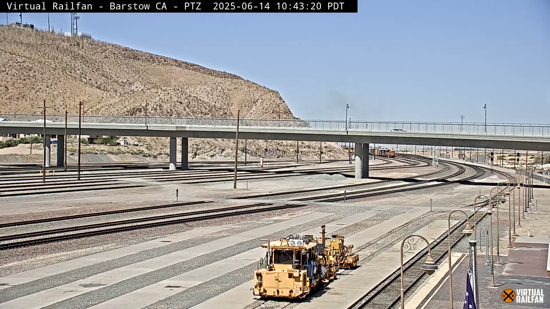 A yellow rail maintenance vehicle sits on a set of train tracks under a sunny sky, near a highway overpass and a desert hillside.