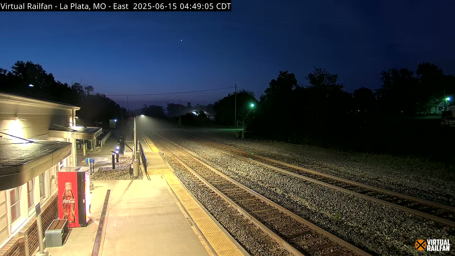 A dimly lit train station platform at night, under a twilight sky, shows multiple train tracks extending into the distance.