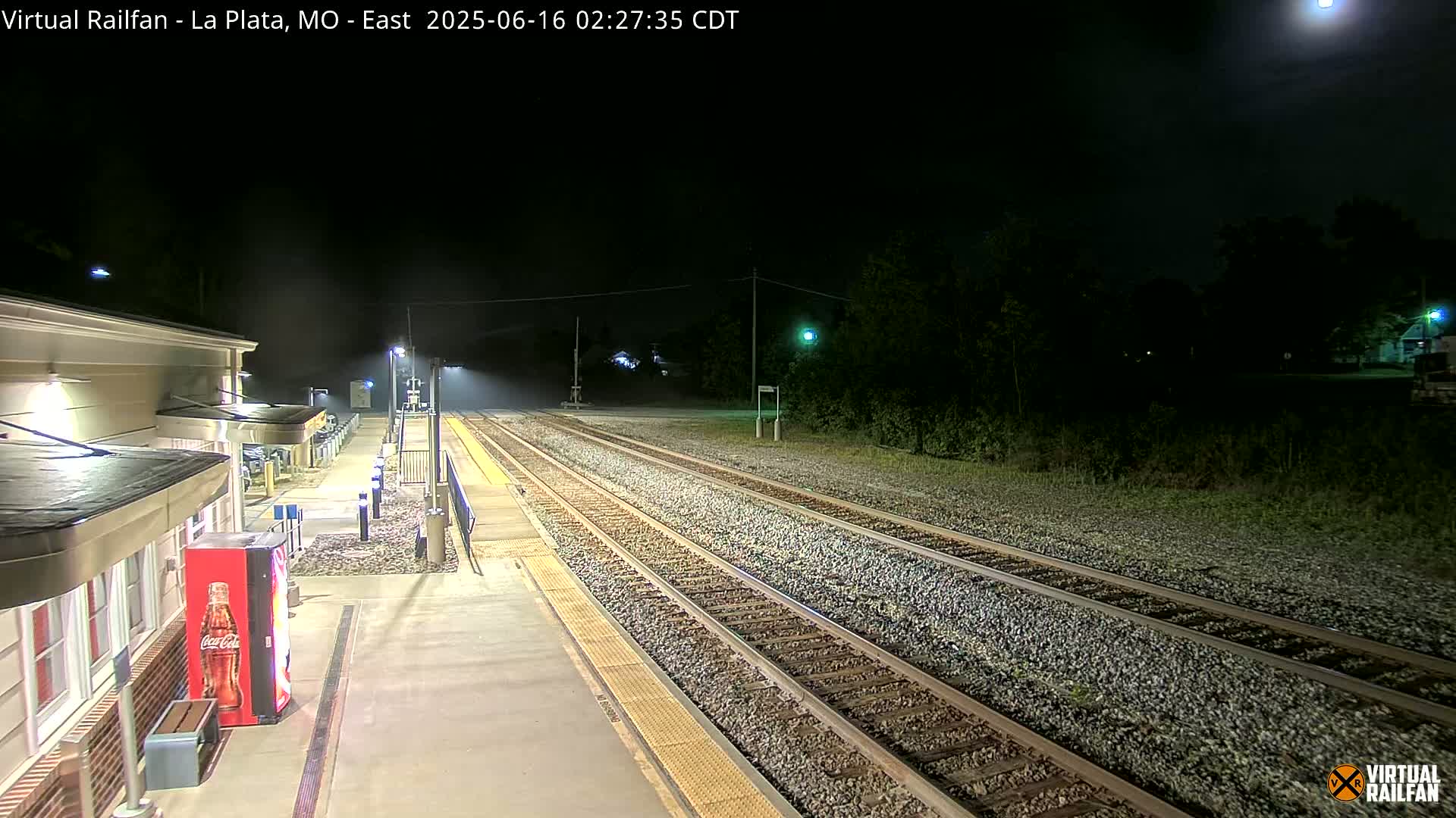 A nighttime view of a train station platform and tracks under a mostly clear sky, illuminated by artificial lights.