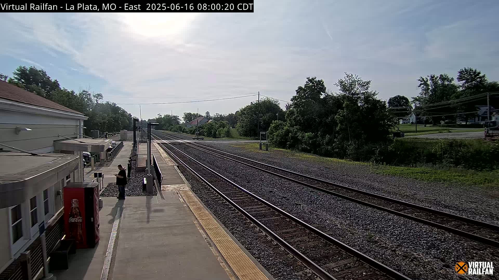 A mostly sunny day at a train station with multiple tracks, a person standing near a Coca-Cola vending machine, and a station building visible in the background.