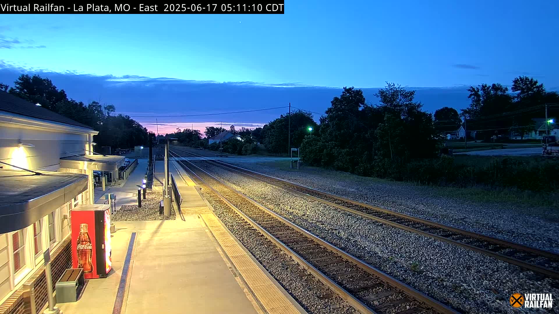 A train station platform at twilight, illuminated by artificial lights, with multiple train tracks extending into the distance under a mostly clear, dark blue sky.
