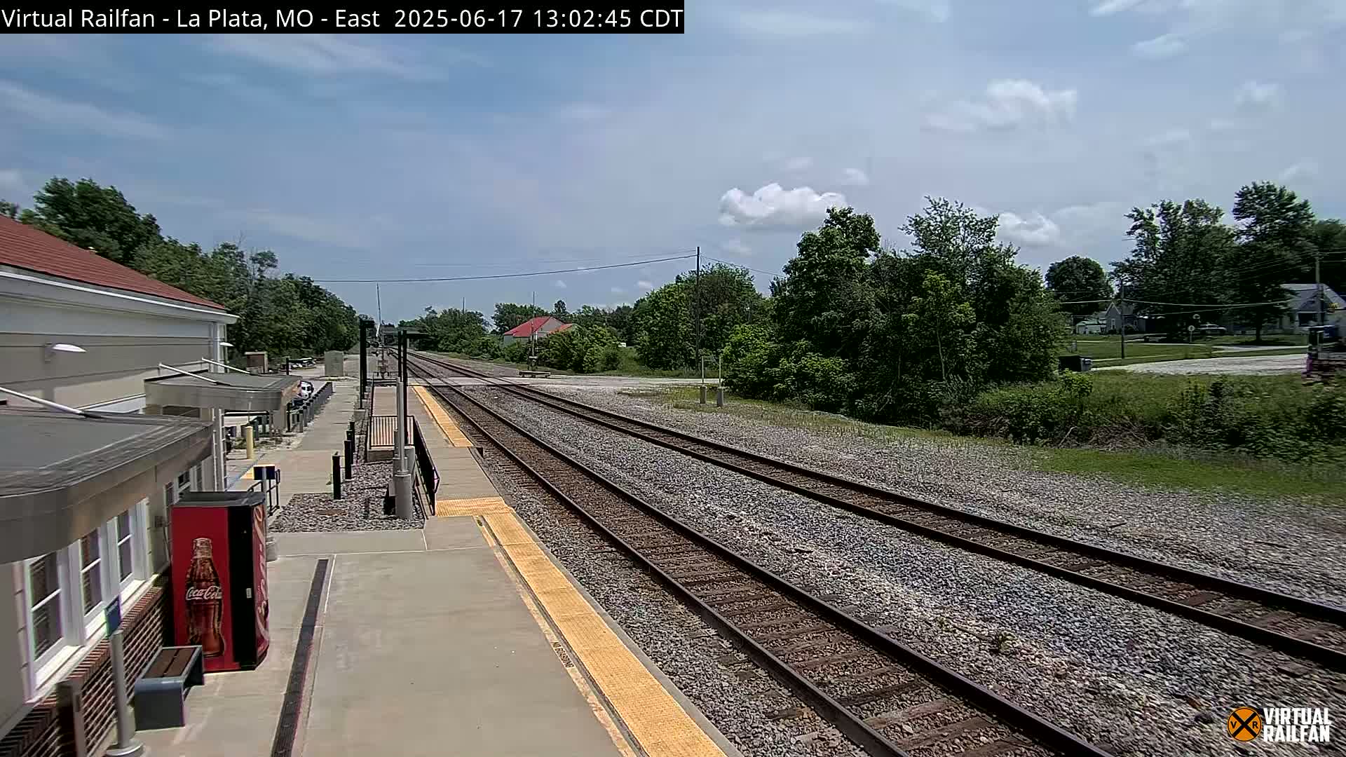 A train station platform with multiple train tracks extending into the distance under a partly cloudy sky.