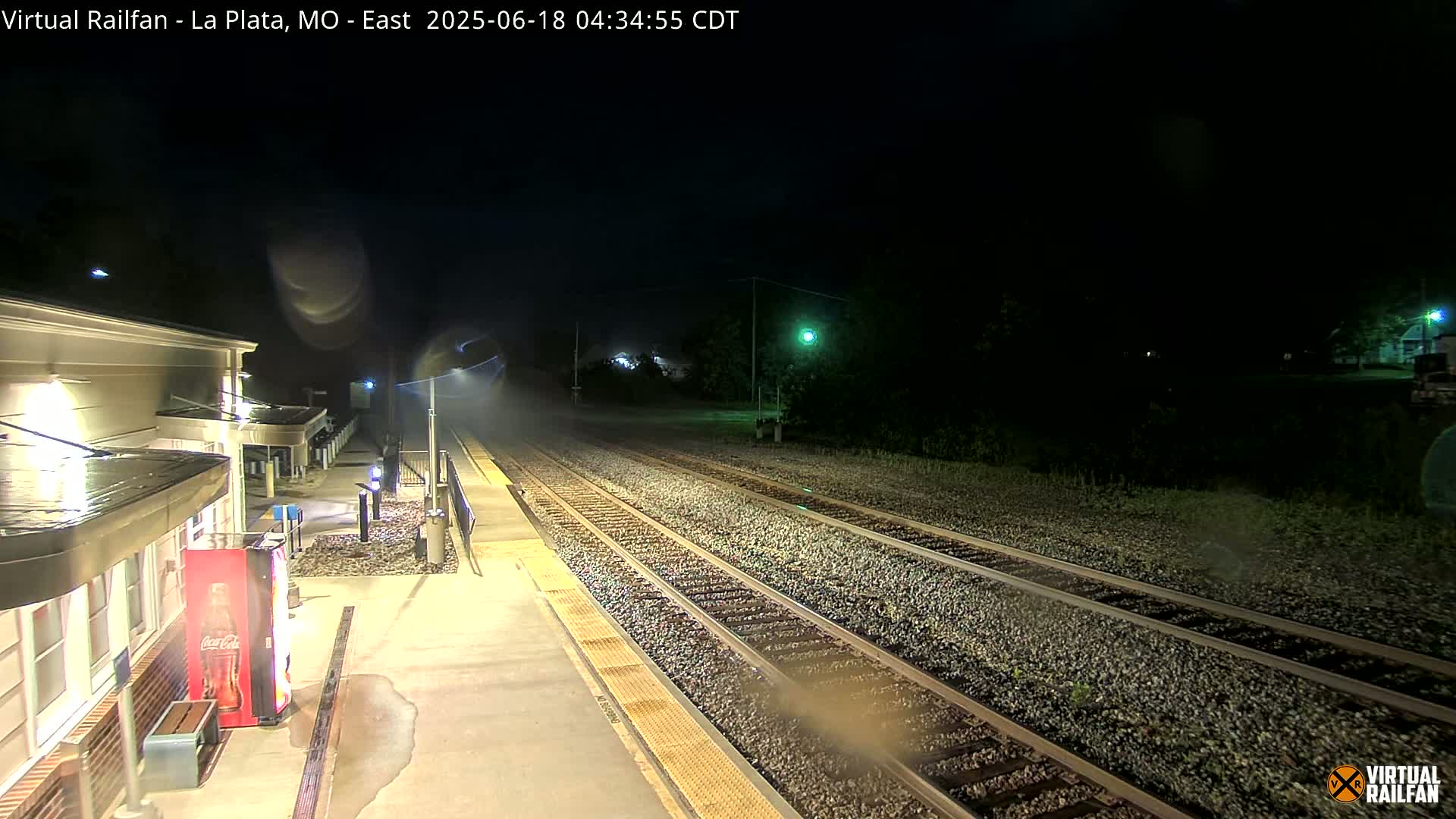 A nighttime view of a rain-slicked train platform and tracks, illuminated by artificial lights.