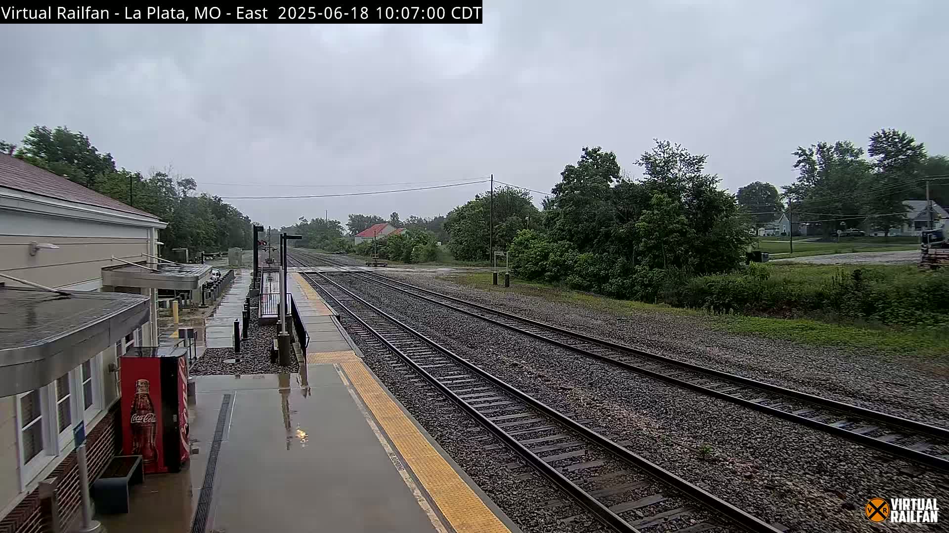 A rain-soaked train station platform with multiple train tracks extending into a tree-lined landscape under an overcast sky.