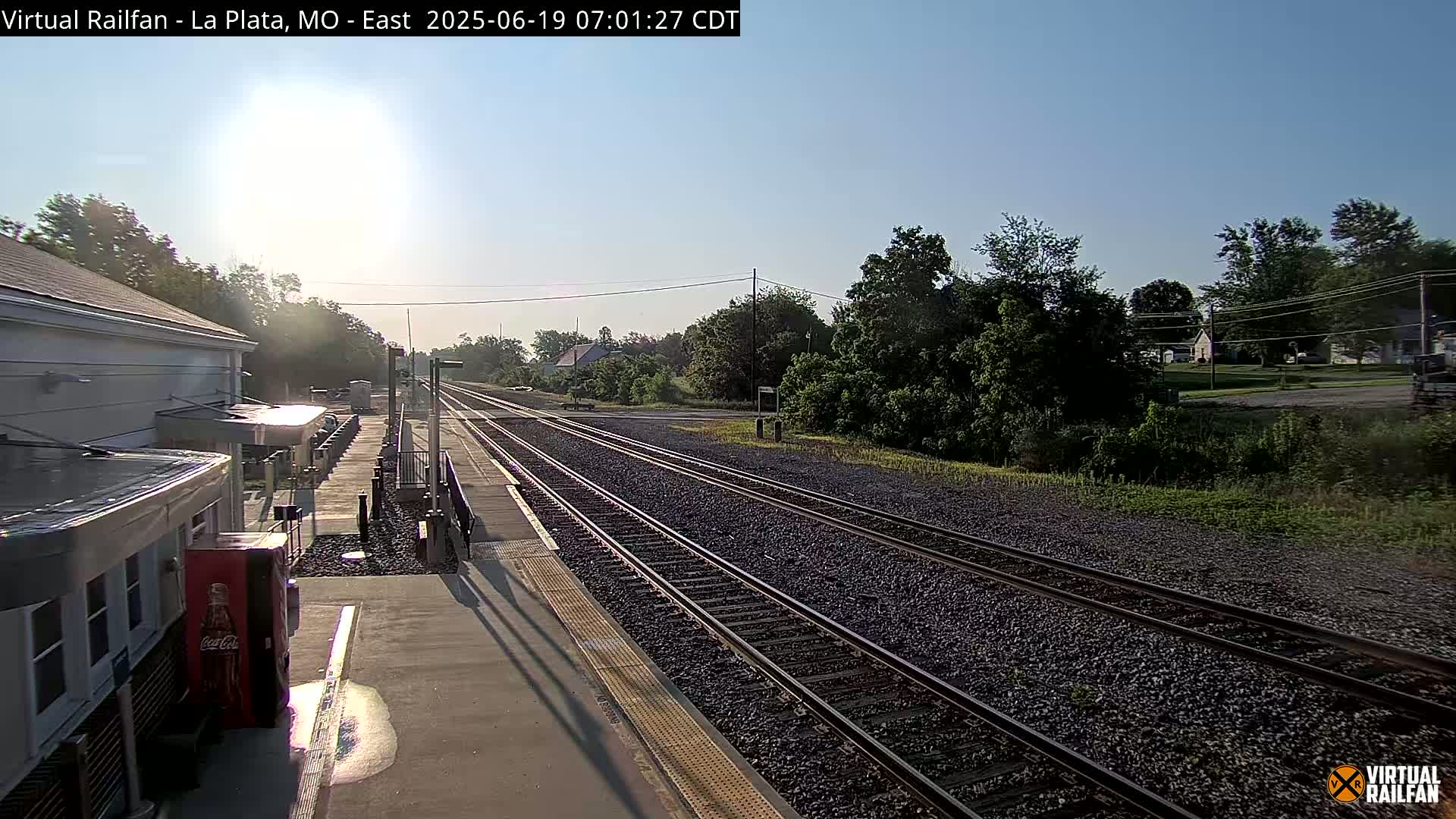 A sunny morning view of a train station platform with two sets of train tracks extending into the distance, bordered by lush green vegetation.
