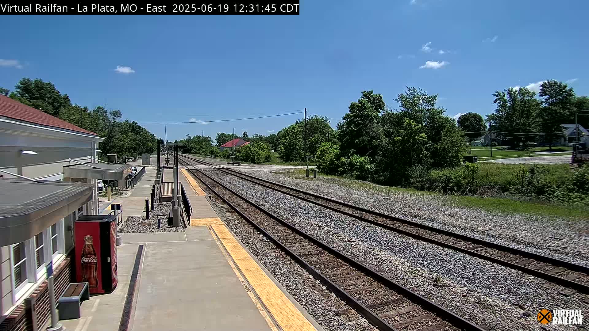 A train station platform with a Coca-Cola vending machine next to it, overlooking a set of train tracks extending into a sunny landscape with lush green trees under a mostly clear blue sky.