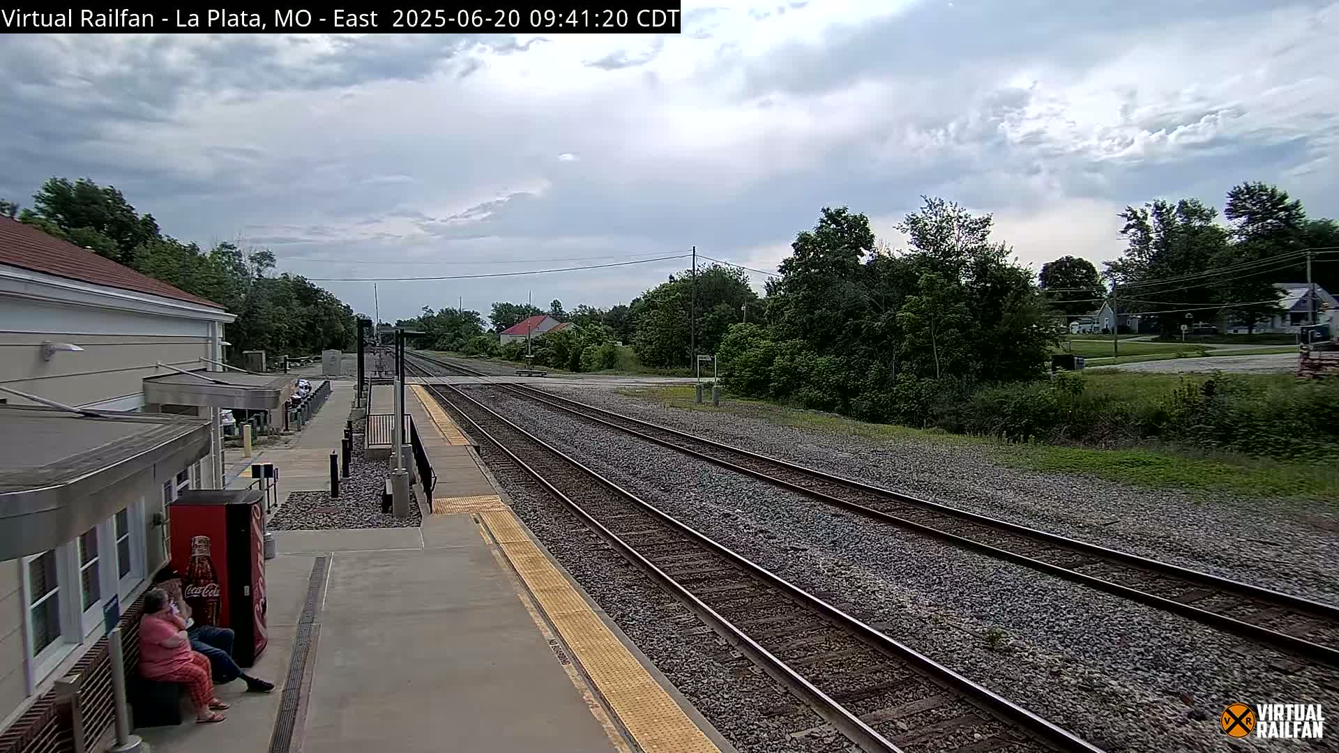 A mostly cloudy day at a train station with multiple train tracks, a station building, and a few people sitting on a bench.