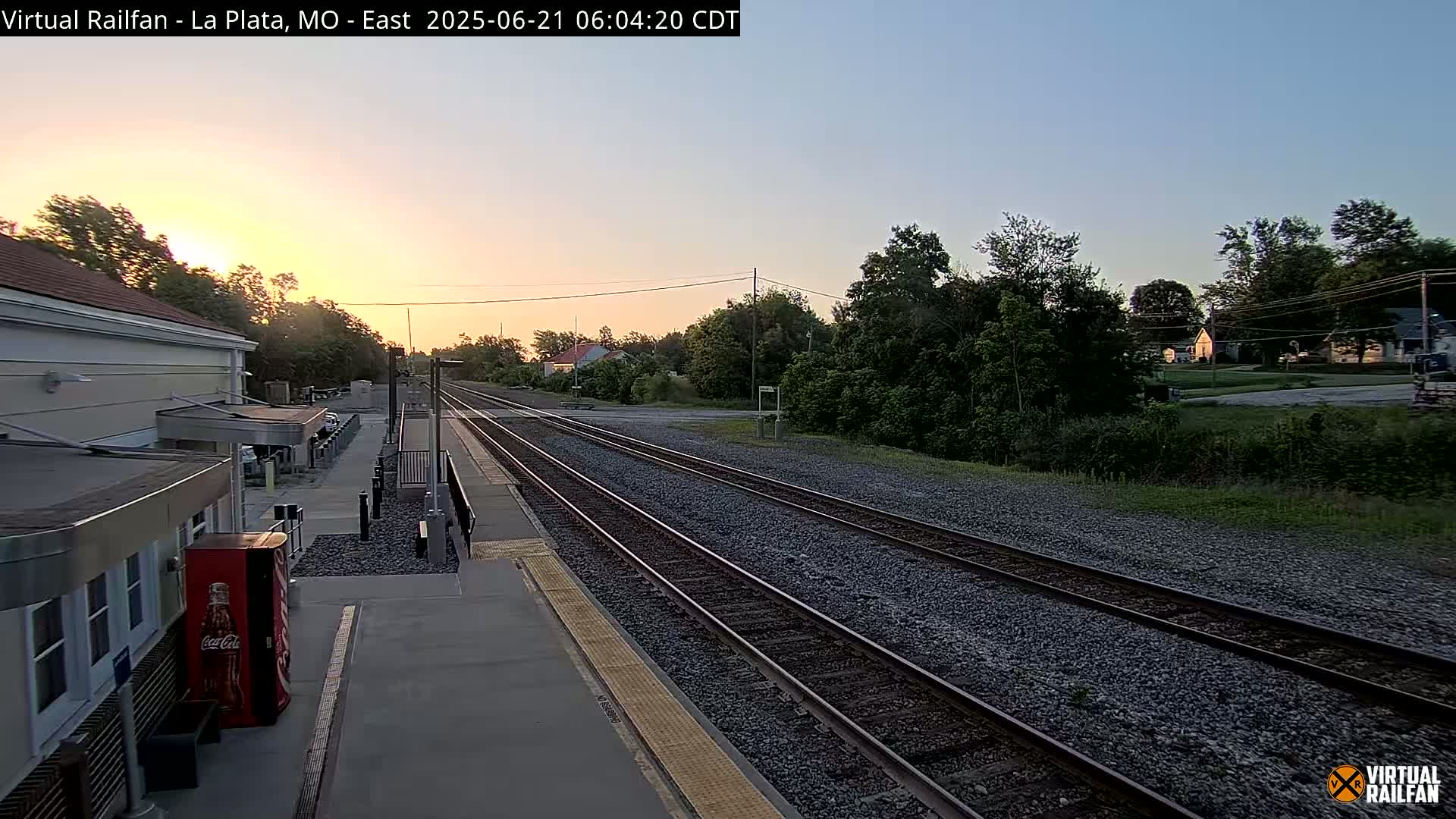 A train station platform at sunrise, with train tracks extending into the distance, is surrounded by trees and houses.