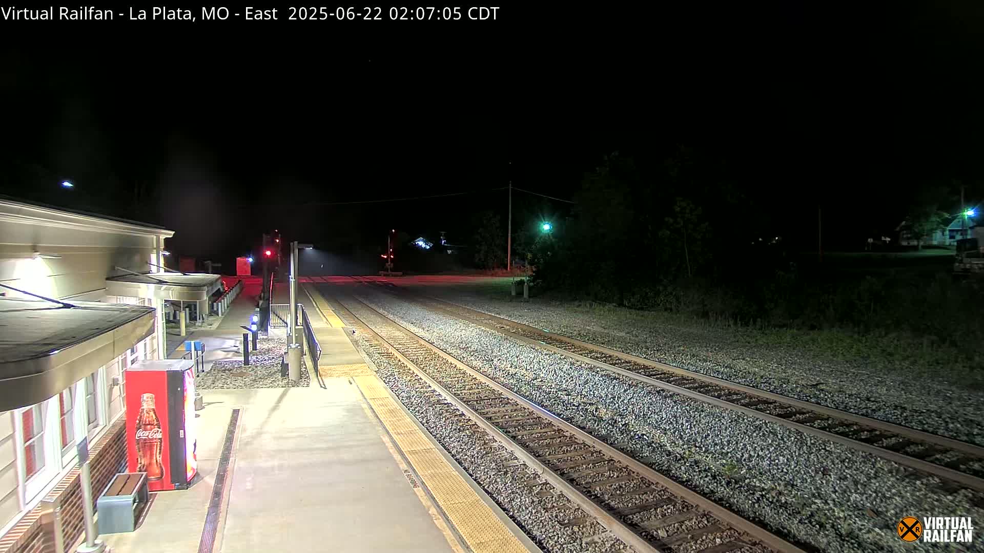 A nighttime view of a train station platform and tracks extending into the dark, with some vegetation visible beyond the tracks.