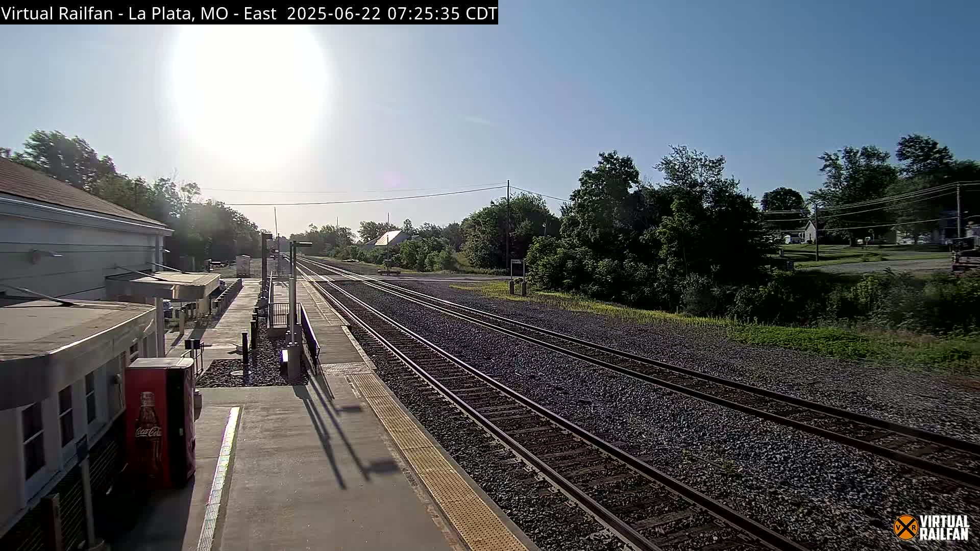A sunny day at a train station with multiple tracks extending into a tree-lined landscape.