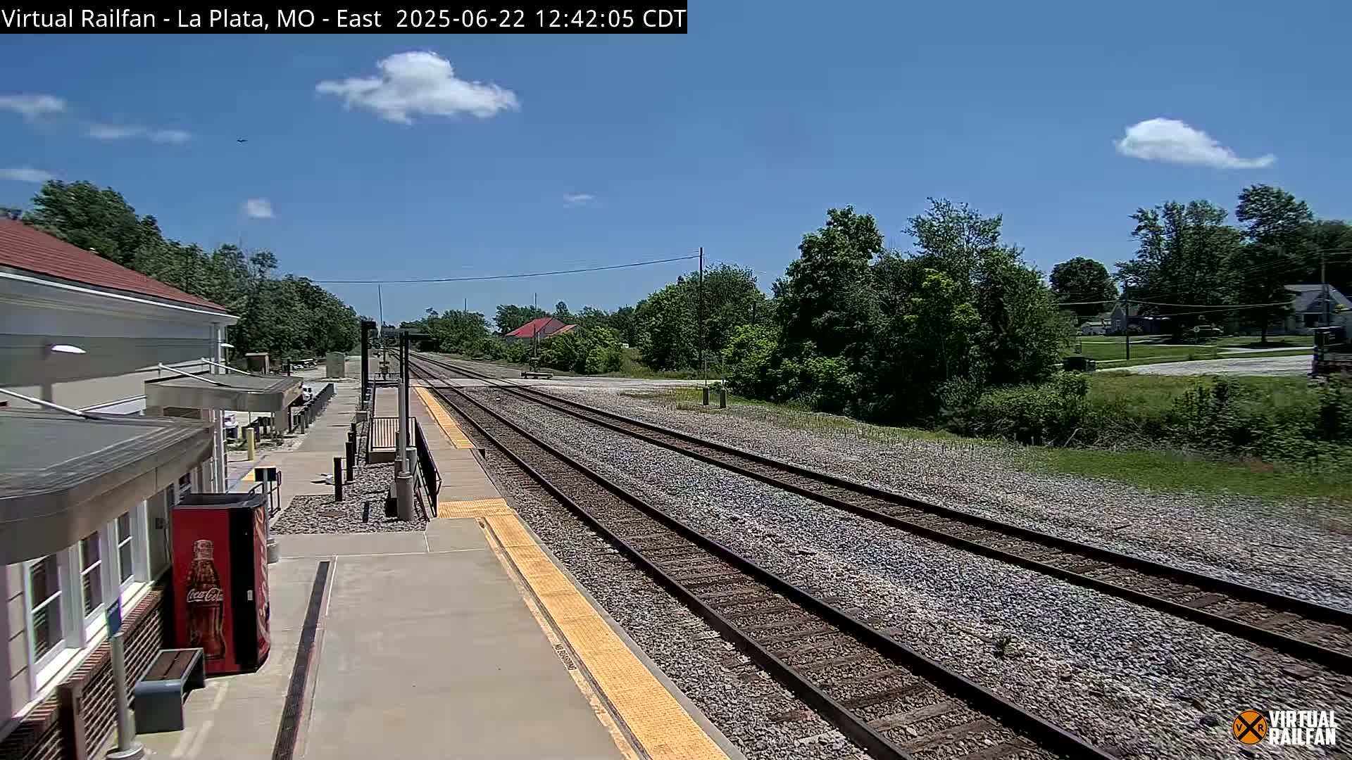 A train station platform with a building and a Coca-Cola vending machine is visible, with train tracks extending into the distance under a partly cloudy sky.