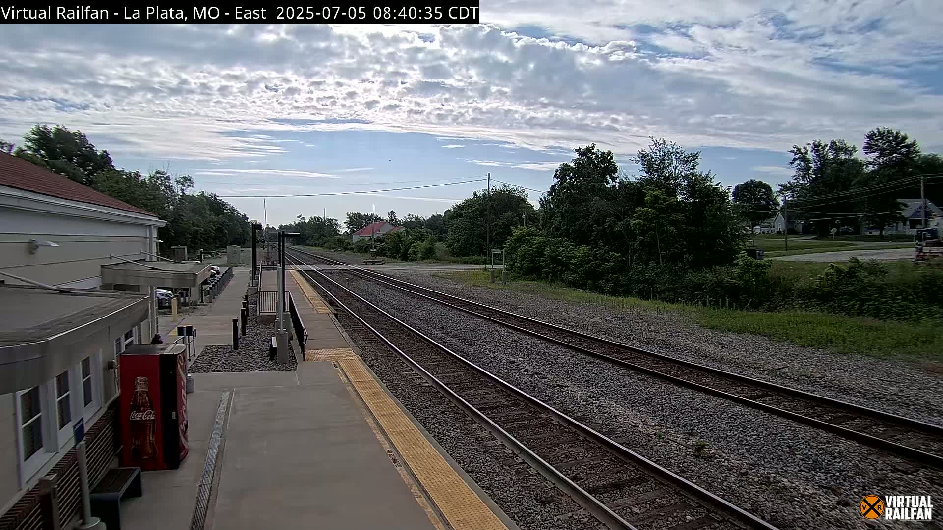 A train station platform with multiple train tracks extends into a partly cloudy landscape.