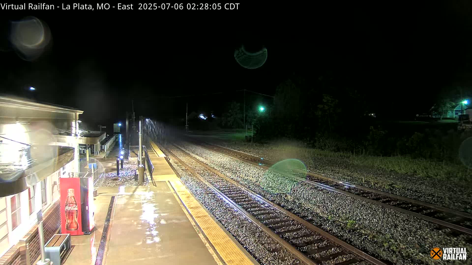 A rain-slicked train platform at night, with train tracks extending into the darkness.