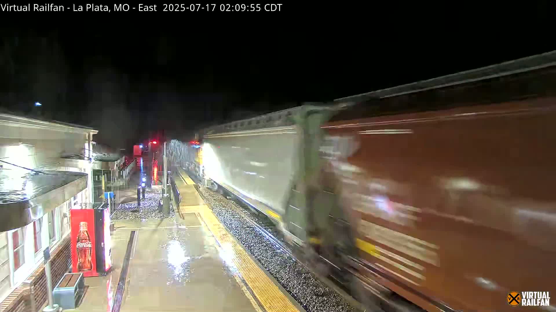 A long freight train passes a rain-soaked train platform at night.