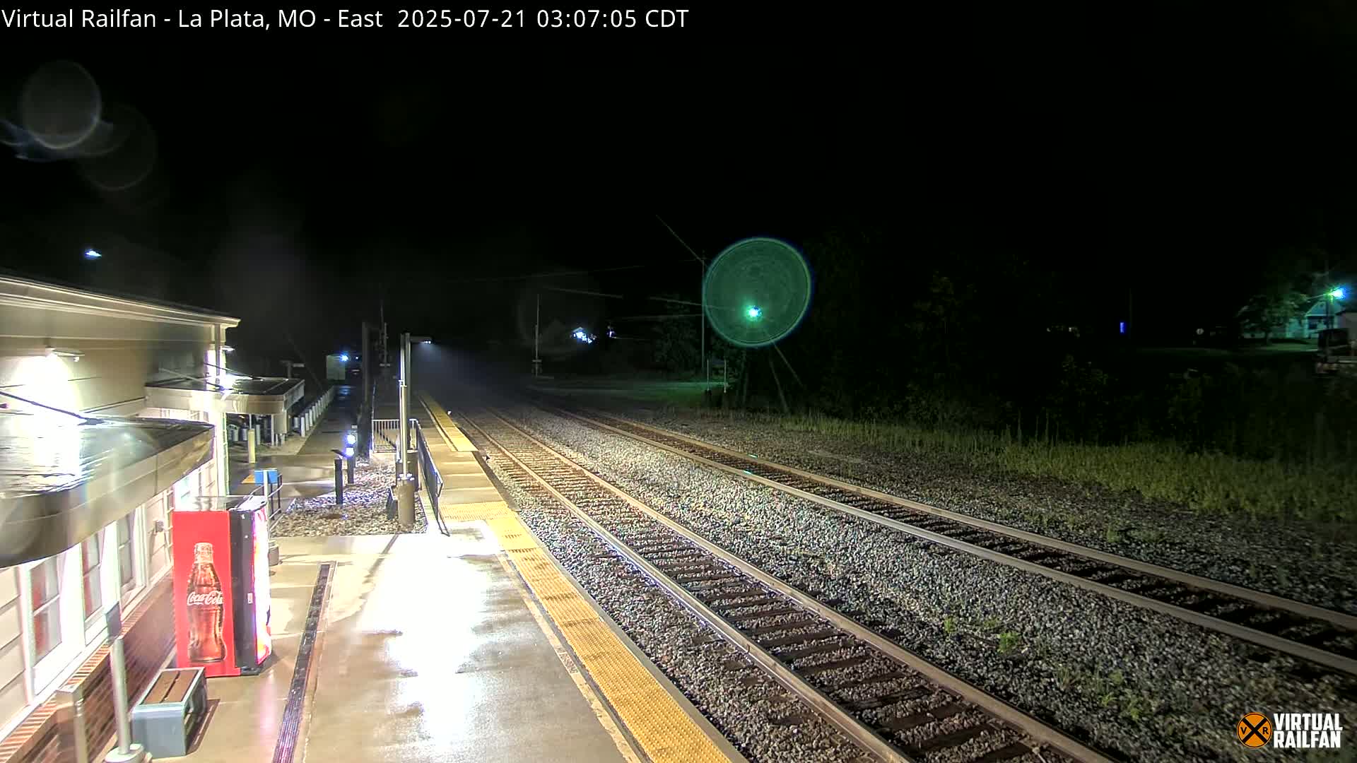 A nighttime view of a wet train platform and tracks, illuminated by artificial lights.