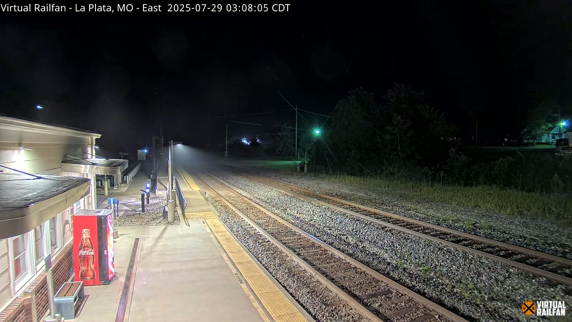 A nighttime view of a train station platform and tracks, with a light mist or fog in the air.