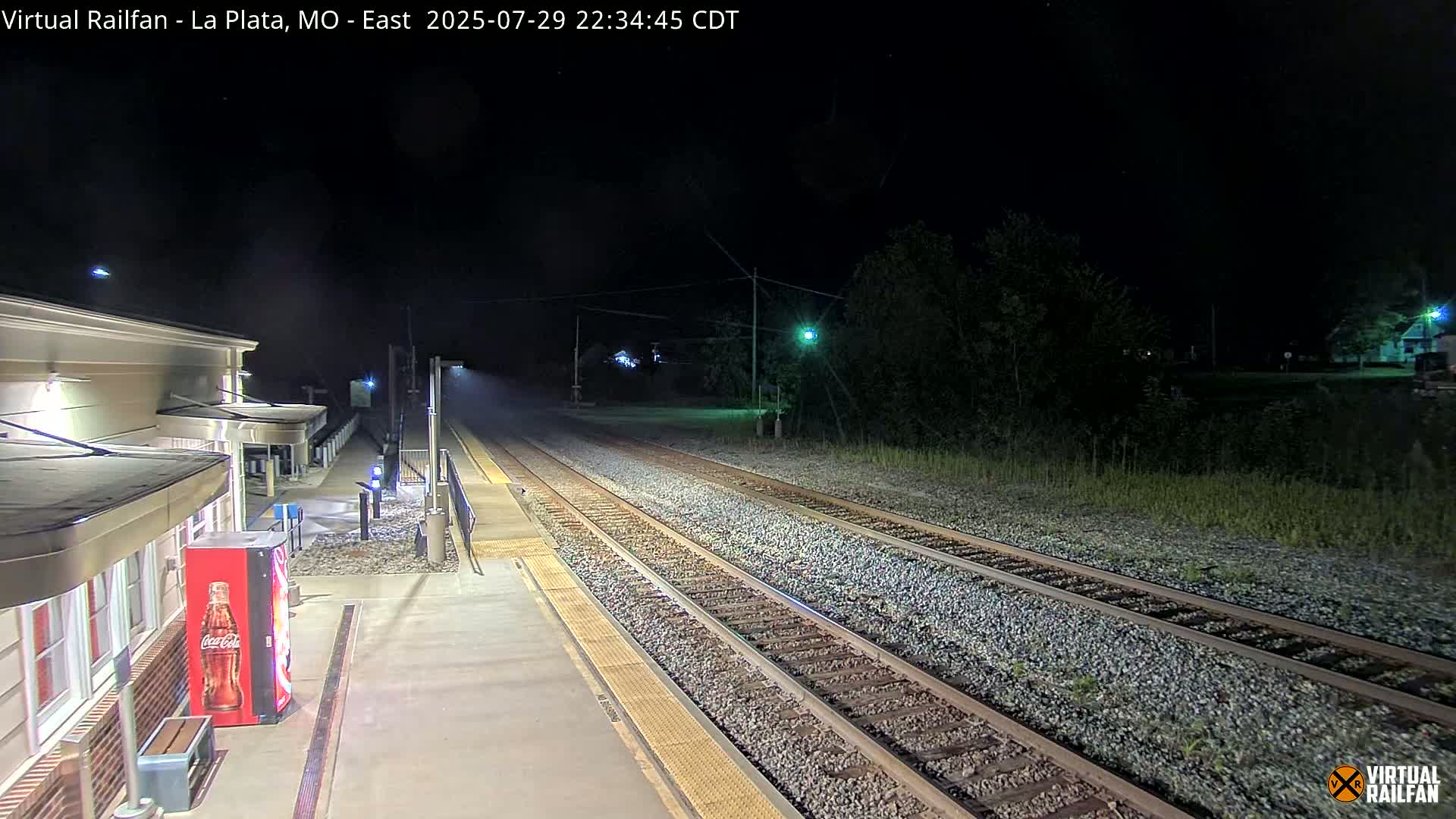 A nighttime view of a train station platform and tracks, with a light mist or fog in the air.