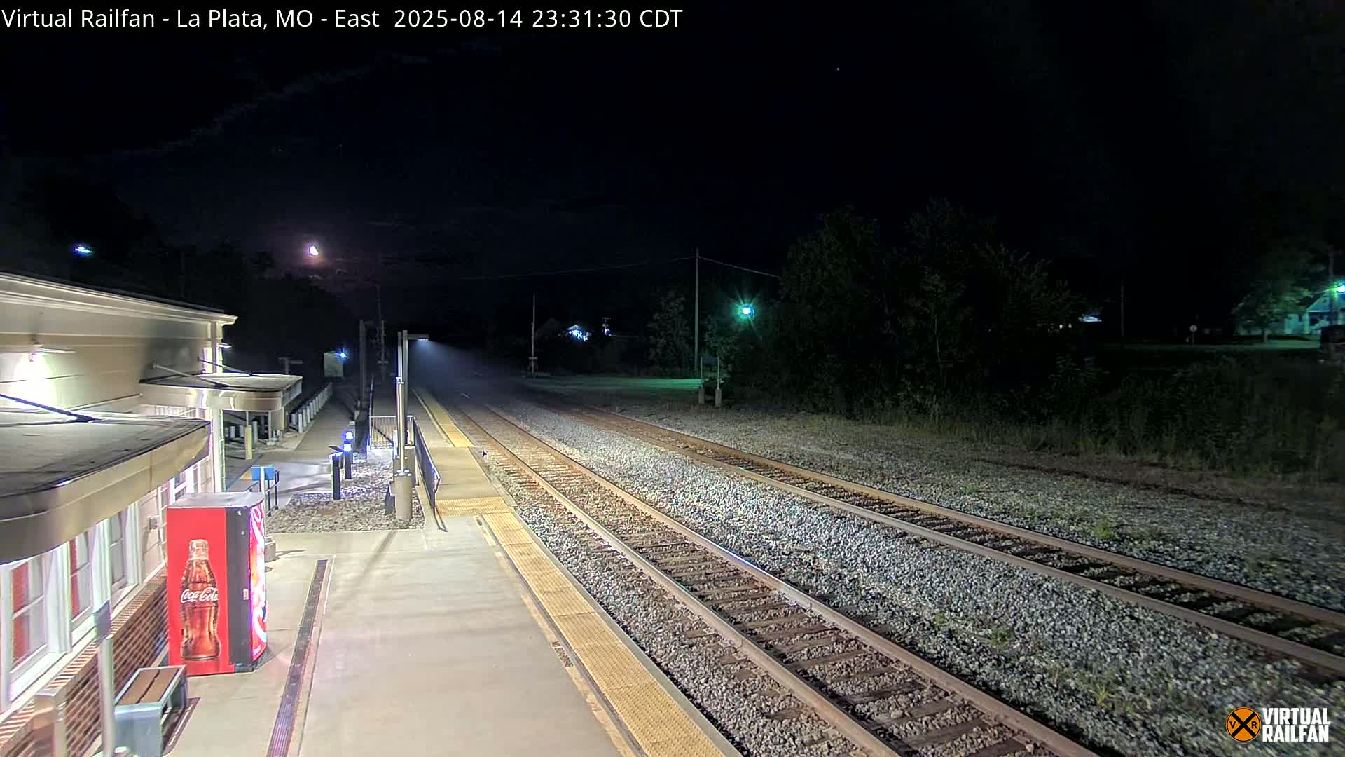 A nighttime view of a train station platform and tracks, with a light mist or fog in the air.