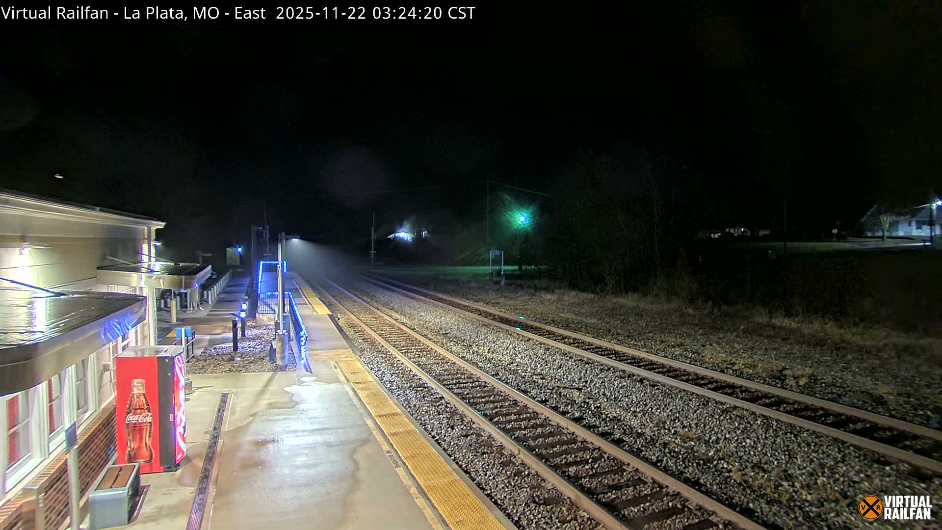 A misty night view shows a wet train station platform with blue-lit railings and multiple tracks receding into the darkness, illuminated by station lights and a distant green signal.