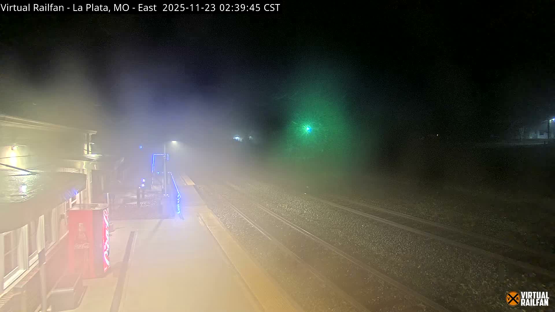 The image captures a foggy nighttime view of a train station platform and railroad tracks, illuminated by various lights including a distant prominent green signal, extending into the haze.