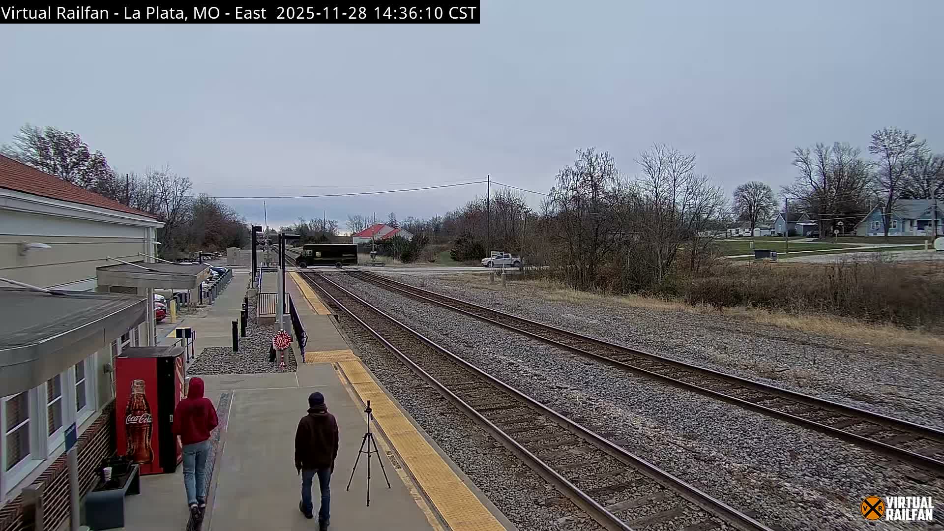 Two people walk along a train station platform adjacent to railroad tracks, with bare trees, distant houses, and a few vehicles visible under an overcast sky on a cool day.