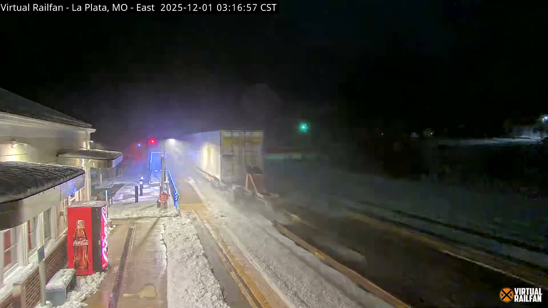 A train passes through a snow-covered station platform at night, illuminated by artificial lights and kicking up a cloud of snow from the tracks in the wintry conditions.