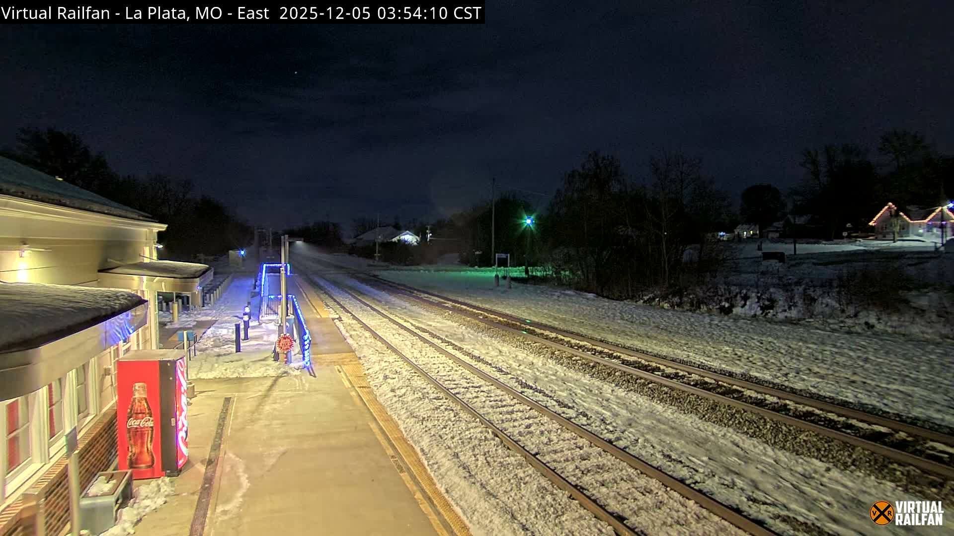 A snowy winter night scene at a train station shows illuminated platforms and tracks covered in snow, with bare trees and distant houses adorned with lights.