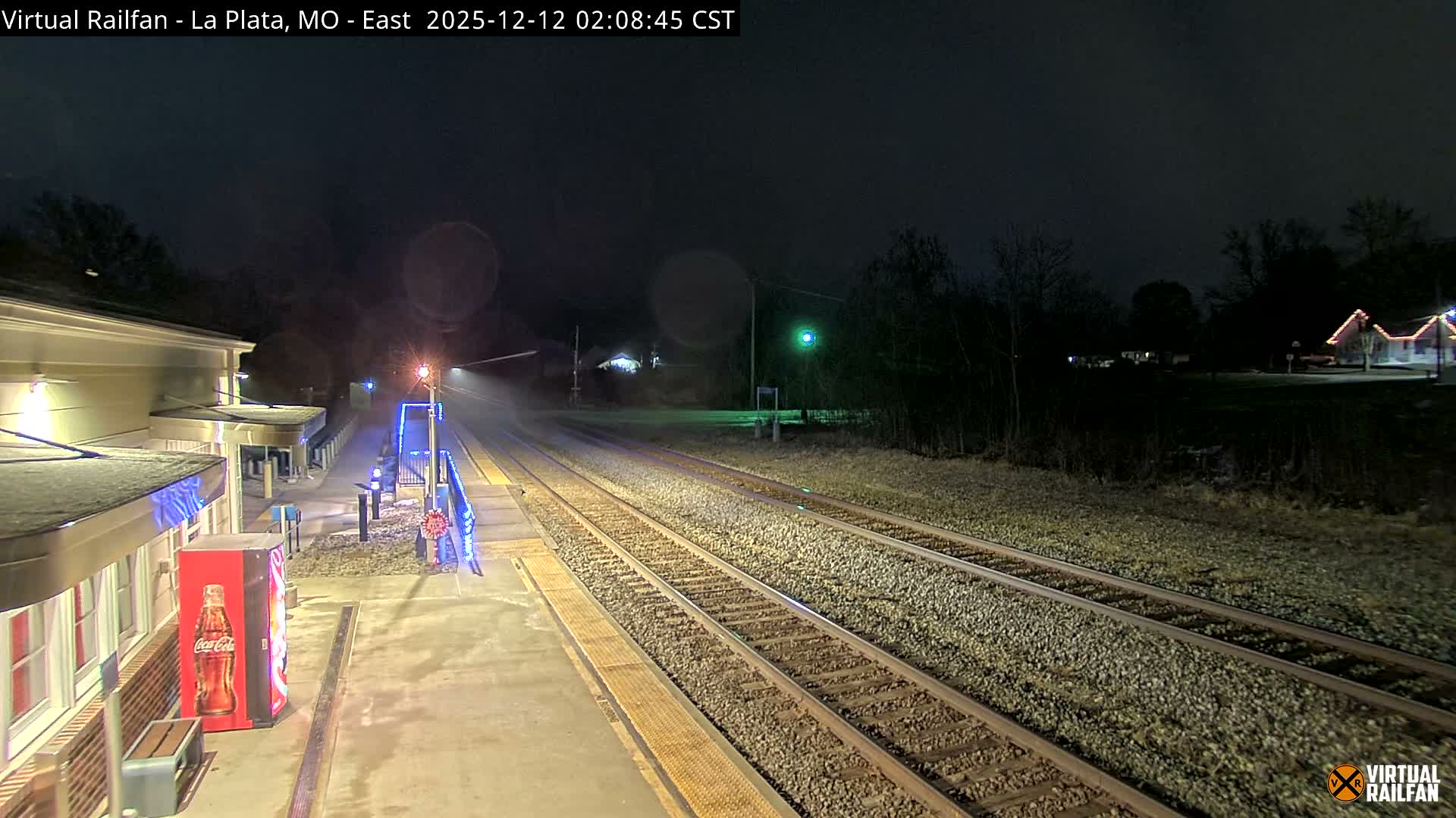 A brightly illuminated train station platform, featuring a red vending machine and blue accent lights, runs parallel to multiple gravel train tracks under a clear, dark night sky, with trees and a few distant lit buildings completing the scene.