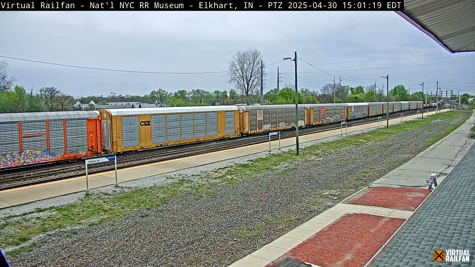 A long freight train, some cars of which are graffitied, sits on a track beside a platform on an overcast day.