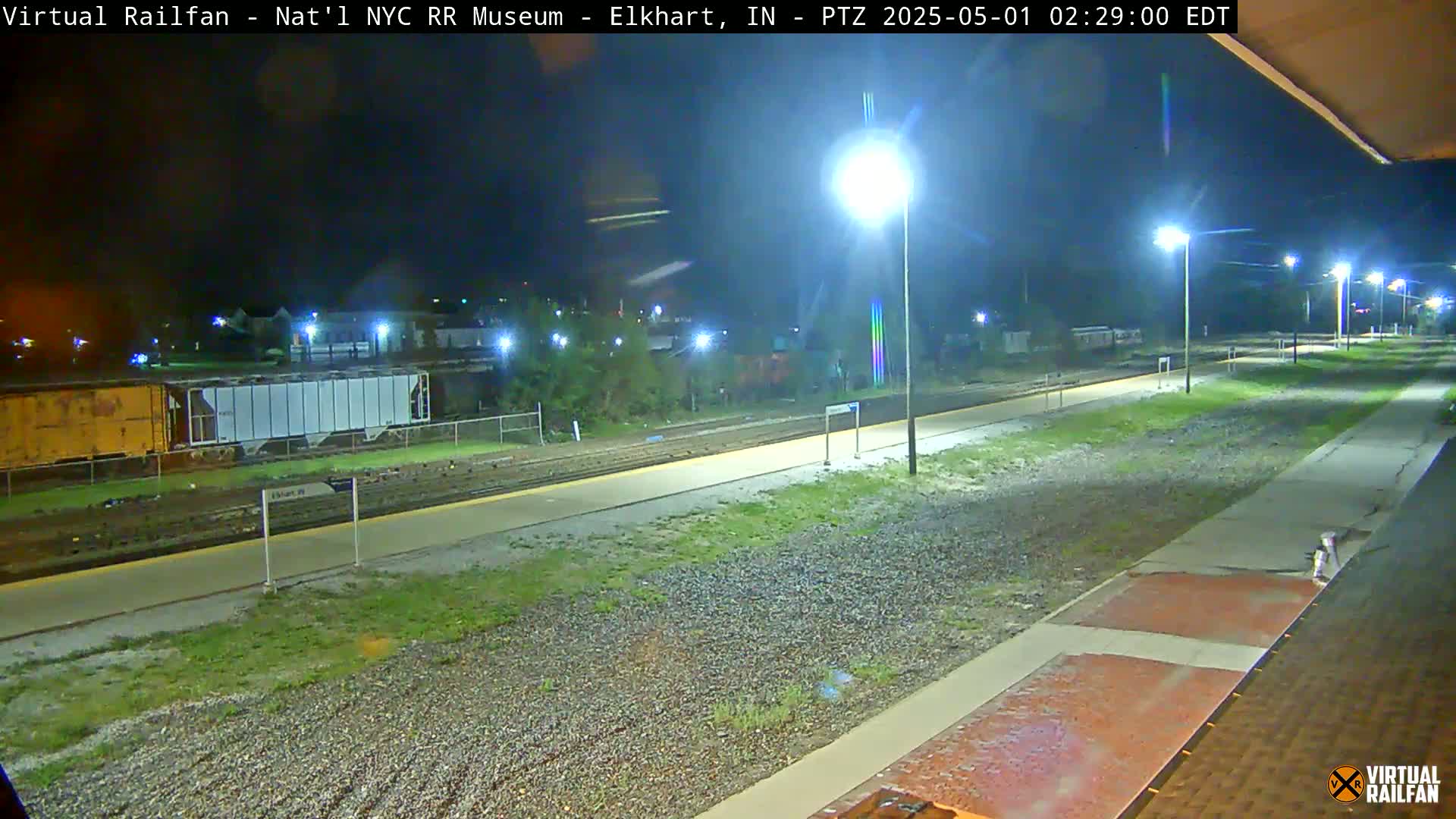 A nighttime view of a train station platform and tracks, illuminated by streetlights, with train cars visible in the background.