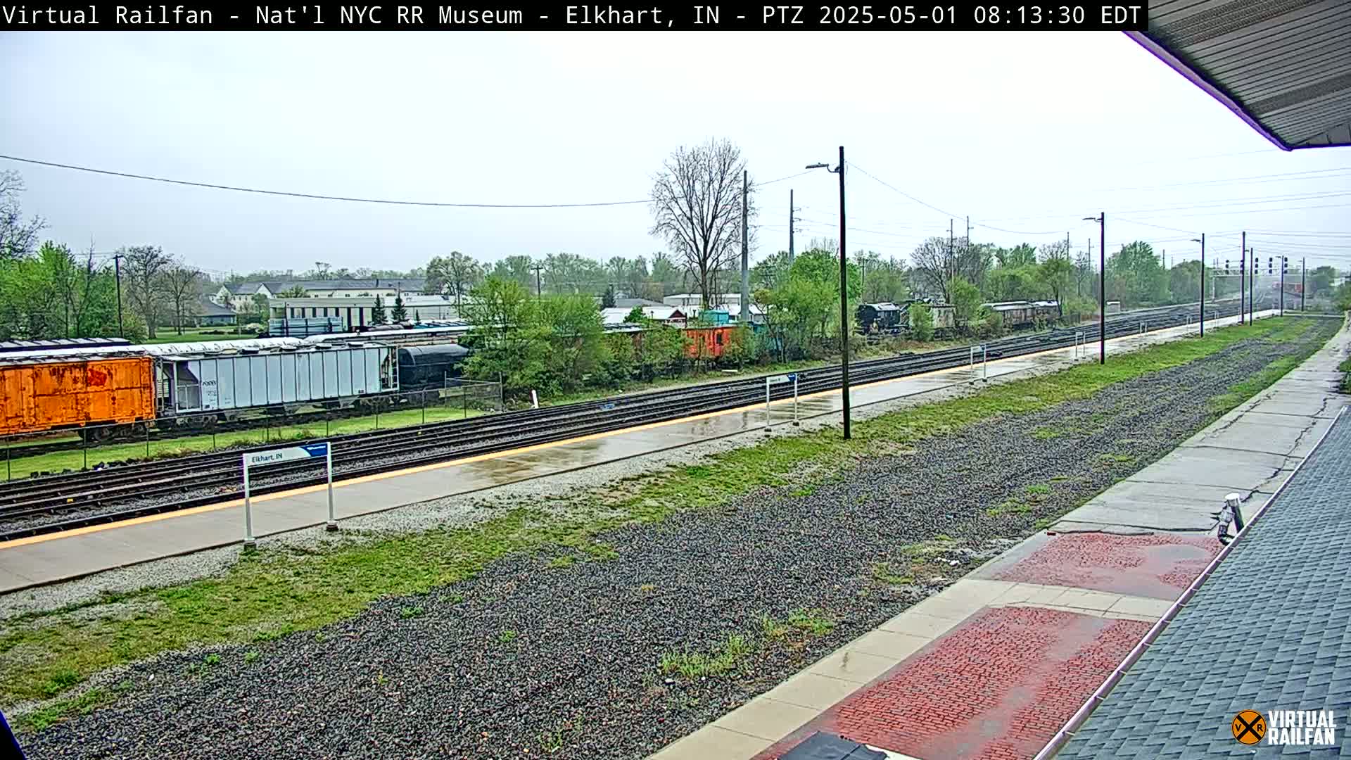 A wet train station platform and several railway tracks, with freight cars and train cars visible in the distance under an overcast sky.