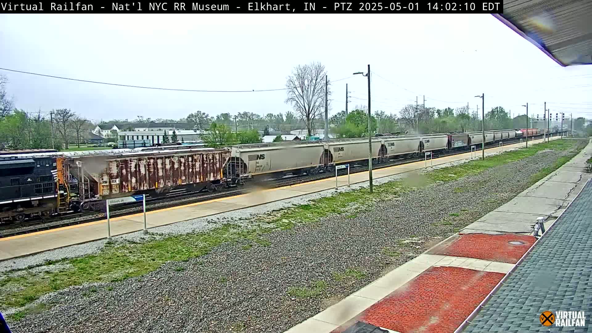 A long freight train, mostly light-gray hopper cars, passes by a station platform on an overcast day.
