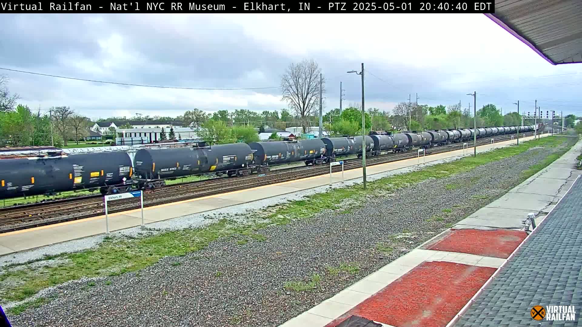 A long freight train of dark-colored tanker cars sits on a track next to a platform under a partly cloudy sky.
