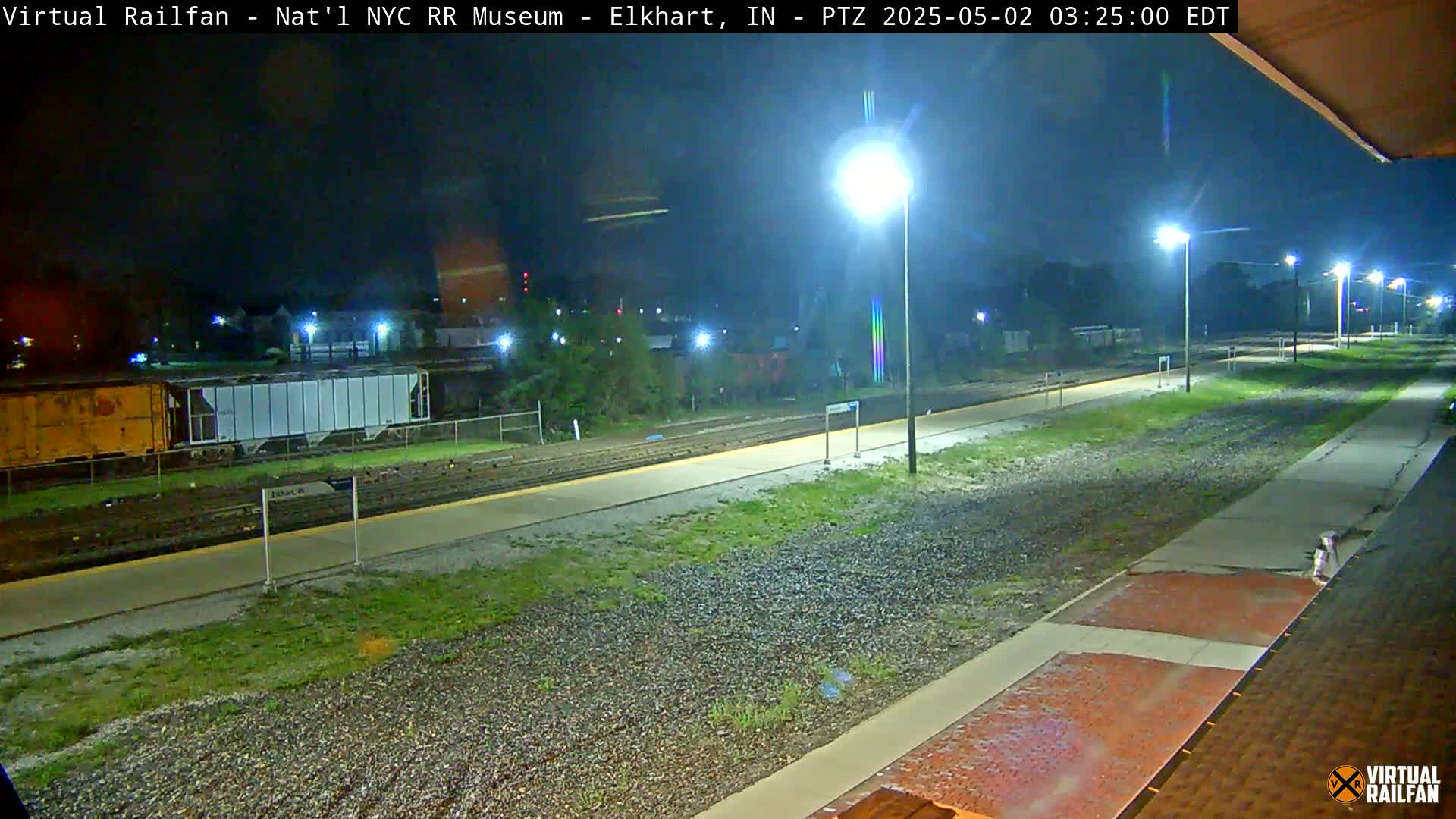 A nighttime view of a train station platform, with several train cars visible in the distance under mostly clear skies.