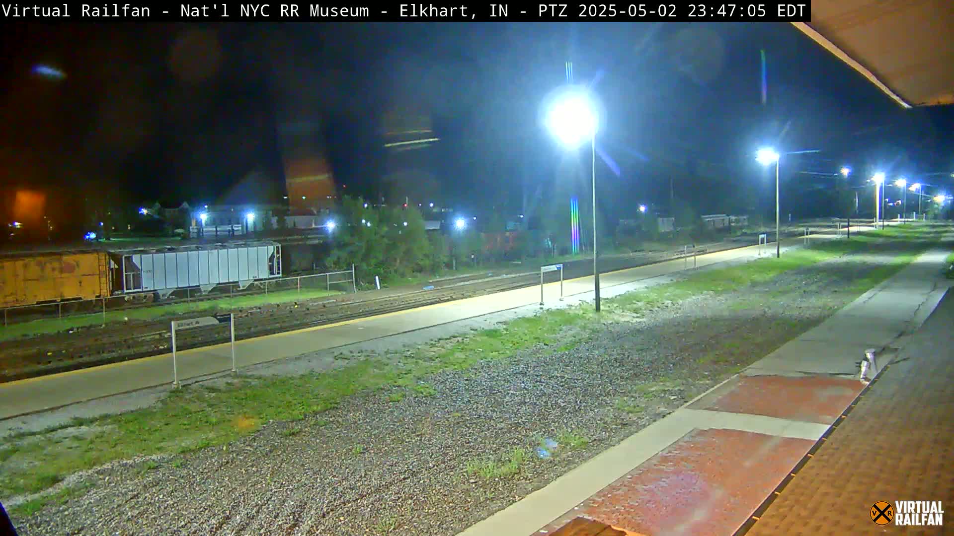 A nighttime view of a train station platform and railroad tracks, illuminated by several bright lights.