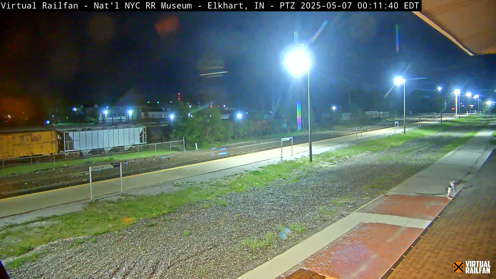 A nighttime view of a railway platform with train cars visible in the distance, illuminated by several streetlights.