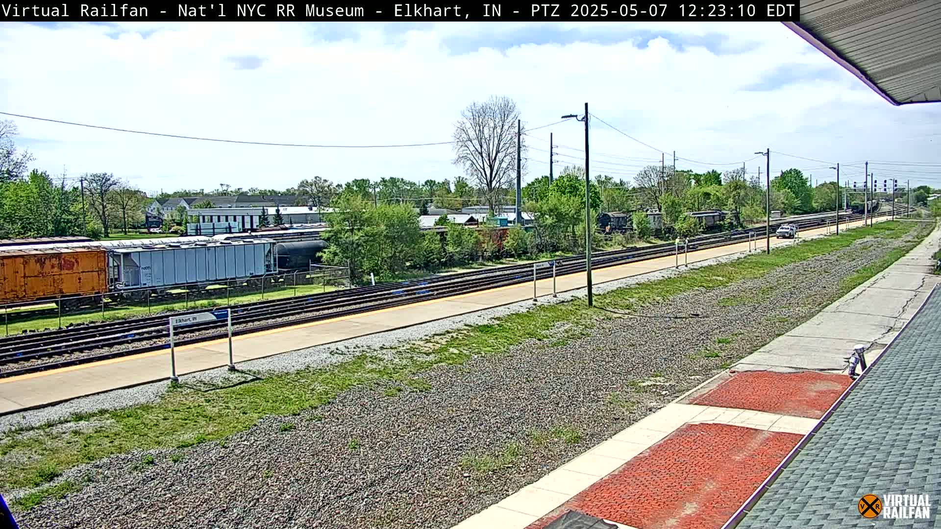 A sunny day shows a train station platform with railway tracks, several freight cars, and a car driving away in the distance.