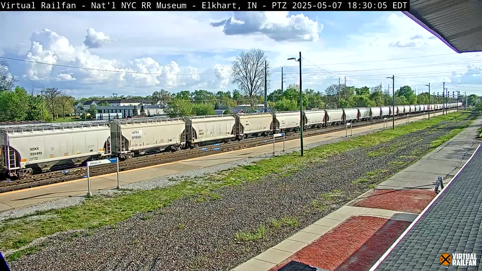 A long freight train of light gray boxcars sits on a railway track under a partly cloudy sky.