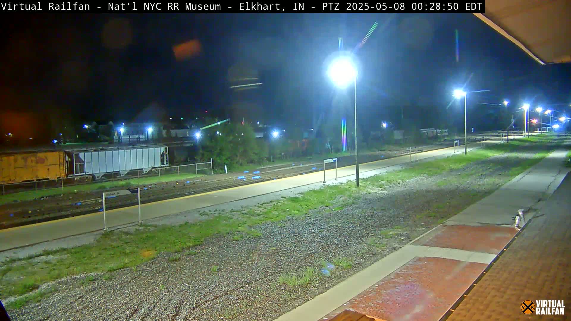 A nighttime view of a train platform and railway tracks, illuminated by streetlights, with several train cars visible in the background.