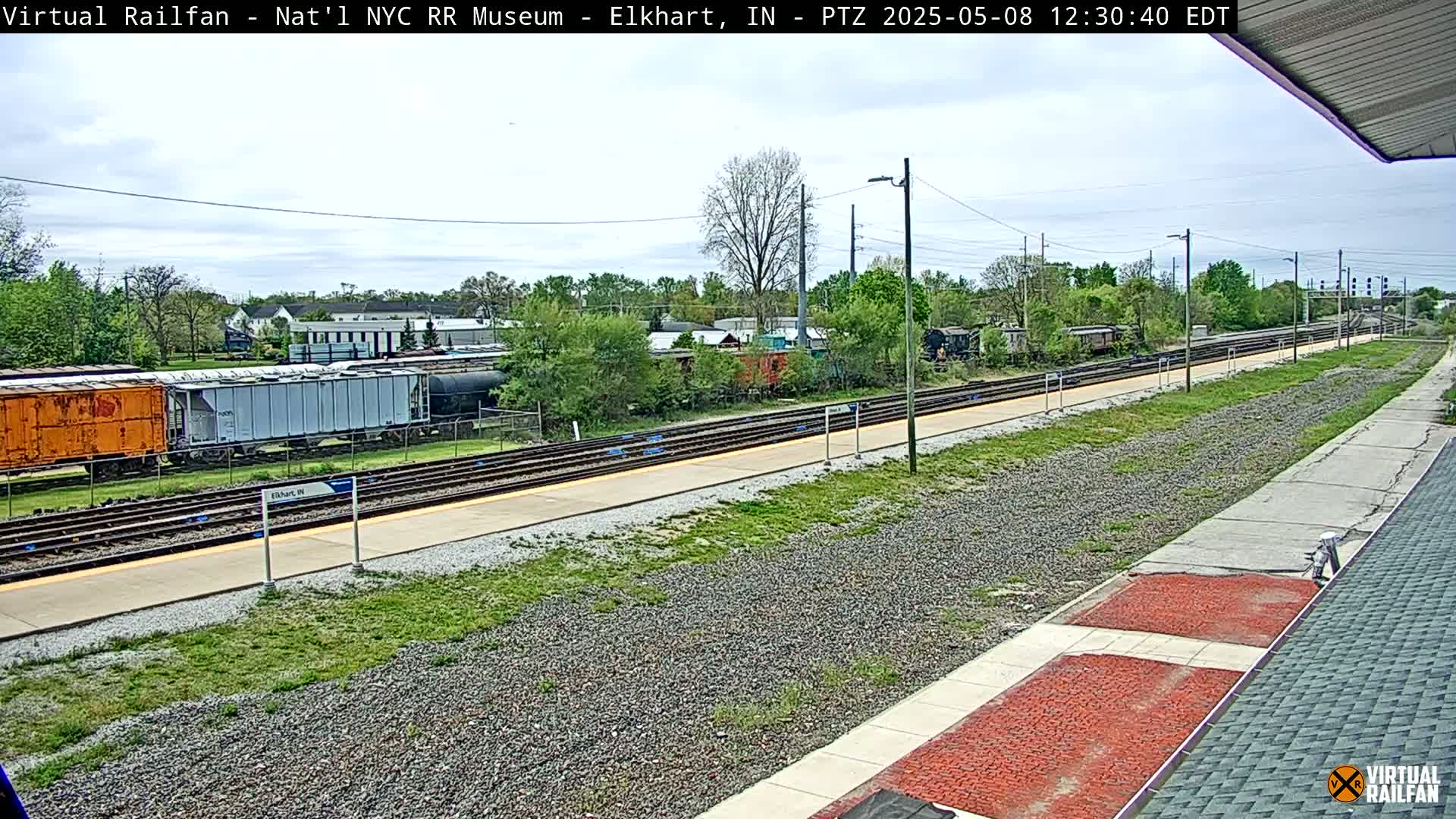 A mostly sunny day shows a train station with several rail cars parked on the tracks, and a paved walkway alongside the tracks, with some grassy areas.