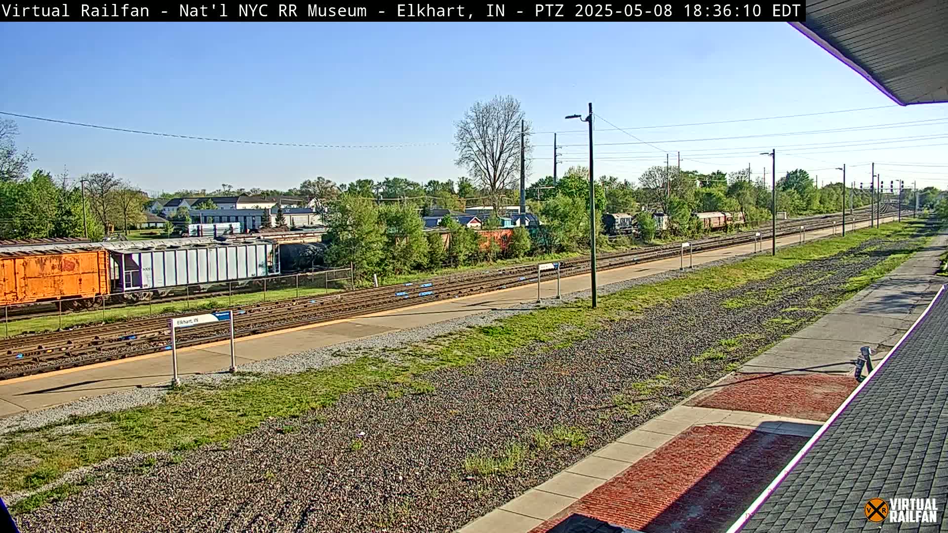 A sunny view of railway tracks and several freight cars, with grassy areas and buildings in the background.