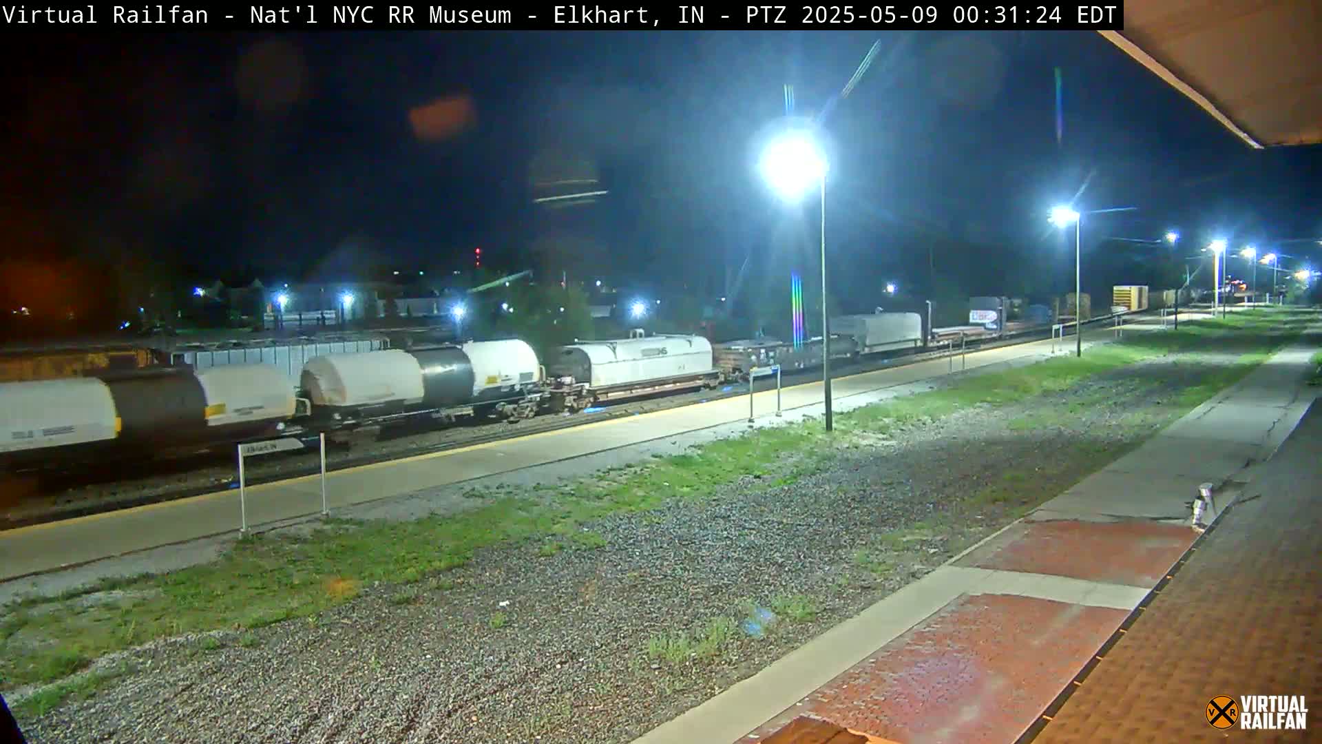 A nighttime view of a train with several tanker cars parked at a station platform, illuminated by several streetlights.